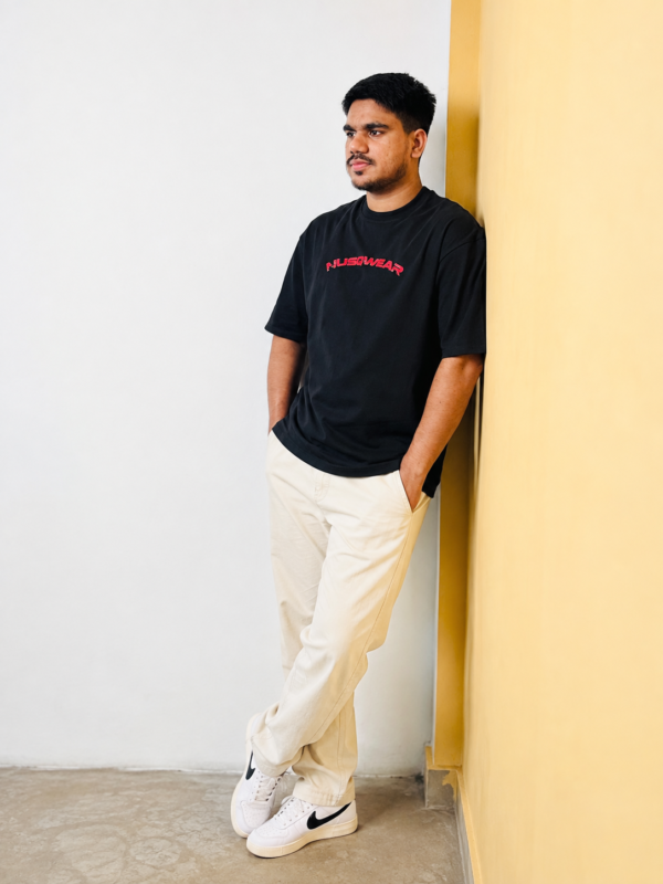 Man wearing black NUSQWEAR T-shirt leaning against yellow wall.